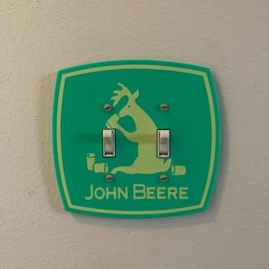 May include: A green light switch cover with a cartoon deer drinking from a cup. The cover has the text "JOHN DEERE" in yellow letters.