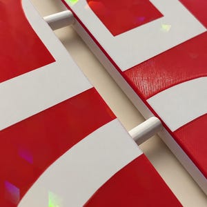 May include: Two red and white panels with a holographic finish featuring geometric shapes. The panels are connected by two white cylindrical rods. The text "Locks together in seconds" is printed at the bottom of the image.