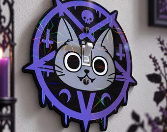May include: A circular light switch cover featuring a grey cat face with large white eyes and a small smile. The design includes a purple pentagram, a skull, and inverted crosses on a black background with a holographic finish and two screws.