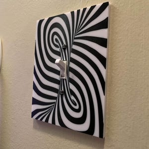 May include: A decorative light switch cover with a black and white optical illusion design. The cover features a swirling pattern of black lines on a white background, with a standard light switch in the center. The switch is in the "ON" position.