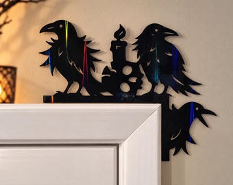 May include: A black, reflective door corner decoration featuring three stylized ravens and a central lit candle. The silhouette design is positioned on the upper right corner of a white door frame, showcasing sharp, angular bird shapes.