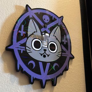 May include: A decorative light switch cover featuring a gray cat face with large white eyes, a small nose, and a smiling mouth. The cat is centered within a purple pentagram design with a skull and crossbones at the top. The switch is in the "ON" position.