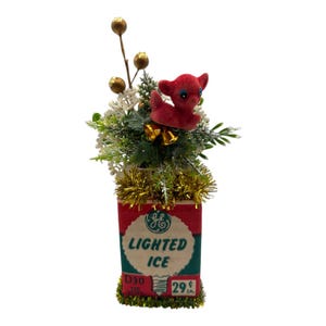 May include: A vintage Christmas decoration featuring a red box with "LIGHTED ICE" text and a red deer figurine. The arrangement includes gold tinsel, faux greenery, and gold bell accents, creating a festive holiday display.