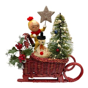 May include: A vintage Christmas decoration featuring a red wicker sled carrying a small Christmas tree, two figurines, and holly. One figurine is holding a glittery star. The sled has red runners and a red woven basket.