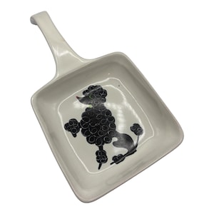 May include: A white ceramic serving dish with a handle, featuring a black poodle silhouette in the center. The dish has a square shape with rounded corners and a raised handle.