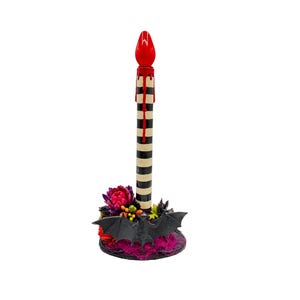 May include: A decorative Halloween candle with a red flame and dripping wax. The candle has a black and white striped body and sits on a base with flowers and a bat. The base is approximately 4 inches in diameter.