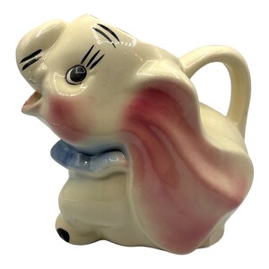 May include: A vintage ceramic pitcher shaped like an elephant with a pink ear, blue bow, and black eyes. The pitcher has a cream-colored base and a pink and white glaze.