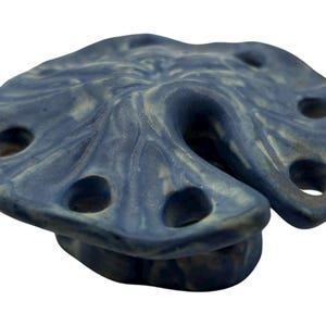 May include: A blue ceramic flower frog with a textured, organic shape resembling a lily pad. The object features a glossy, mottled finish with lighter blue and grey highlights and includes several circular openings for arranging stems.