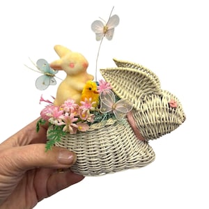 May include: A white wicker bunny-shaped basket filled with pink flowers, a yellow bunny, a yellow chick, and decorative butterflies. The basket has a pink ribbon and is held by a hand.