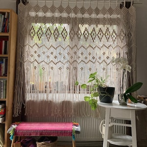 May include: A macrame window curtain with a diamond pattern hangs in front of a window. The curtain is beige and has a fringe at the bottom. A small white table holds a potted plant and an orchid. A bench with a pink woven top sits below the window.