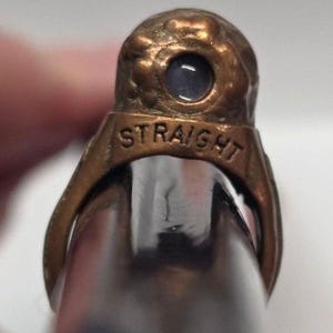 May include: A bronze-toned metal ring featuring a textured, rounded top with a small, embedded blue circular stone. The band of the ring is clearly engraved with the word STRAIGHT in capital letters, positioned directly below the stone setting.