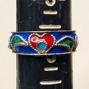 May include: A silver-toned ring with a blue enamel band. The ring features a red heart with a blue center, flanked by green leaf-shaped designs. The ring is on a black cylindrical object.