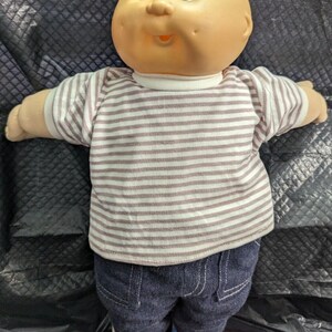 May include: A doll with a round, bald head, dimpled cheeks, and blue eyes. The doll wears a short-sleeved shirt with thin horizontal brown and white stripes, dark blue denim pants with visible stitching, and white sneakers with blue accents.