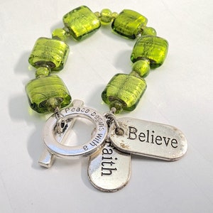 May include: A green beaded bracelet with square and round beads. The bracelet has silver charms that read "Believe", "faith", and "peace begins with a". The bracelet is on a white surface.