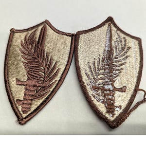 May include: Two shield-shaped embroidered patches. Each patch features a brown sword and a fern-like design on a tan background, framed by a brown border. The patches are connected and appear to be new.