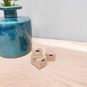 Könnte beinhalten: Eine türkisfarbene Keramikvase mit breitem Sockel und schmalem Hals, die grüne Pflanzenstiele enthält. Drei kleine, braune Pappkartons sind auf einer Holzoberfläche gestapelt. Jede Box hat schwarze Griffe und das Wort "Wichtelumzug" aufgedruckt.