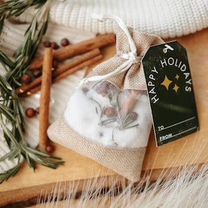 May include: A small burlap bag tied with a string, containing a white, snow-like substance with sprigs of rosemary and cinnamon sticks. A black tag with "Happy Holidays" is attached. The scene includes a candle and a cozy, neutral-toned aesthetic.