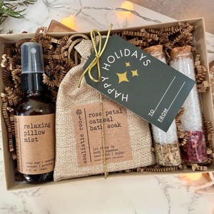 May include: A gift box with self-care items. It includes a brown glass bottle of pillow mist, a burlap bag of bath soak, and two glass tubes with bath salts. A tag reads "Happy Holidays".