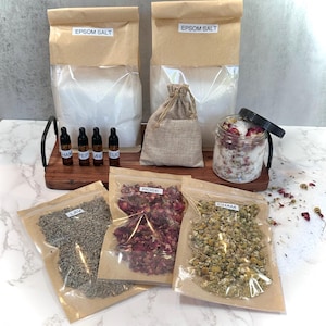 May include: A spa-themed collection featuring Epsom salt in paper bags, small essential oil bottles, and dried herbs in clear bags. A glass jar with bath salts and rose petals is also included. The labels read: Epsom Salt, HAM, PEP, AV, EUC, LAV, ROSE, and CHAM.