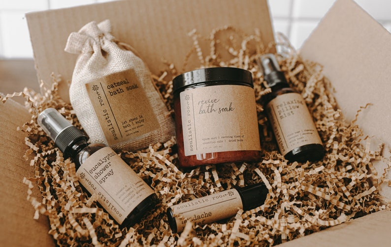 May include: A cardboard box filled with brown crinkle paper and a variety of bath and body products. The products include a small burlap bag labeled "oatmeal bath soak", a glass bottle labeled "eucalyptus shower spray", a glass jar labeled "revive bath soak", a small glass bottle labeled "relaxing pillow mist", and a small roller bottle labeled "headache".