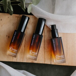 May include: Three amber-colored glass bottles of facial serum with black pumps and caps. The bottles are labeled "Soulistic Root" and "Renew Facial Serum." The bottles are arranged on a wooden surface with greenery and white fabric.