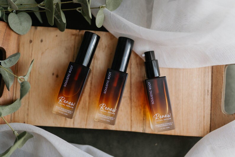 May include: Three amber-colored glass bottles of facial serum with black pumps and caps. The bottles are labeled "Soulistic Root" and "Renew Facial Serum." The bottles are arranged on a wooden surface with greenery and white fabric.
