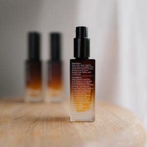 May include: A skincare serum bottle with a black pump and a gradient amber and clear glass body. The label includes directions and ingredients. Two additional bottles are blurred in the background. The bottles are on a wooden surface.