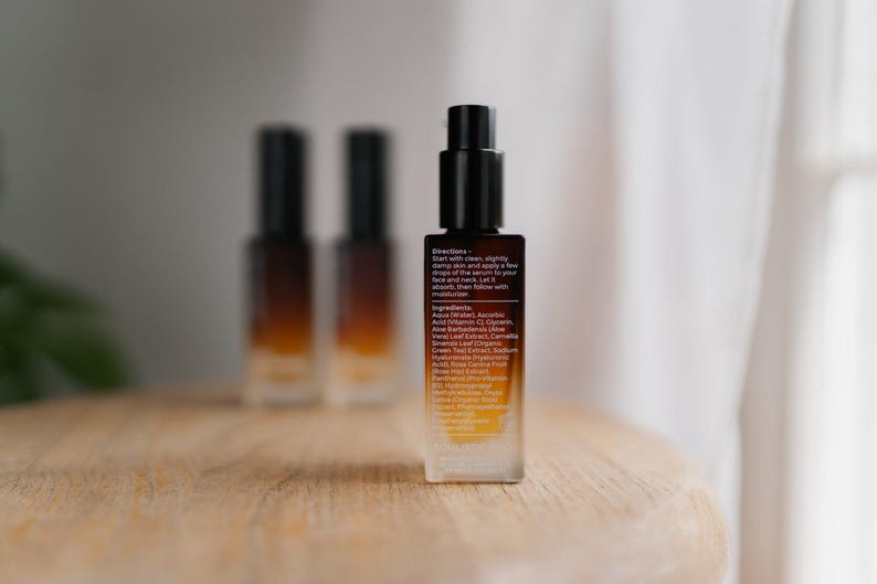 May include: A skincare serum bottle with a black pump and a gradient amber and clear glass body. The label includes directions and ingredients. Two additional bottles are blurred in the background. The bottles are on a wooden surface.