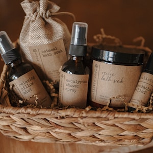 K&ouml;nnte beinhalten: Ein Weidenkorb gef&uuml;llt mit braunem, zerkleinertem Papier und verschiedenen Bade- und K&ouml;rperpflegeprodukten. Die Produkte umfassen eine braune Flasche mit der Aufschrift "Eukalyptus-Duschspray", eine braune Flasche mit der Aufschrift "Entspannendes Kissenspray", eine braune T&uuml;te mit der Aufschrift "Haferflocken-Badesalz", ein Glas mit der Aufschrift "Belebendes Badesalz" und eine kleine braune Flasche mit der Aufschrift "Kopfschmerzmittel".