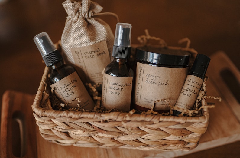K&ouml;nnte beinhalten: Ein Weidenkorb gef&uuml;llt mit braunem, zerkleinertem Papier und verschiedenen Bade- und K&ouml;rperpflegeprodukten. Die Produkte umfassen eine braune Flasche mit der Aufschrift "Eukalyptus-Duschspray", eine braune Flasche mit der Aufschrift "Entspannendes Kissenspray", eine braune T&uuml;te mit der Aufschrift "Haferflocken-Badesalz", ein Glas mit der Aufschrift "Belebendes Badesalz" und eine kleine braune Flasche mit der Aufschrift "Kopfschmerzmittel".