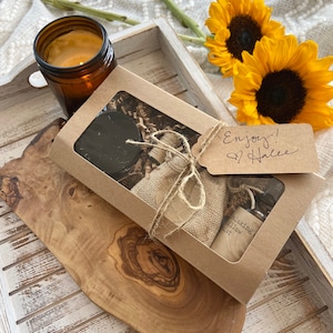 May include: A brown gift box with a clear window, filled with a candle, a small bottle of essential oil, and a burlap bag. The box is tied with twine and a tag that says "Enjoy!  From Hallie".