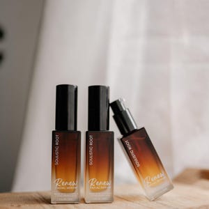 May include: Three bottles of Soulistic Root Renew Facial Serum on a wooden surface. The bottles have a gradient brown and clear design with black caps. The text "Renew Facial Serum" is visible on the bottles.