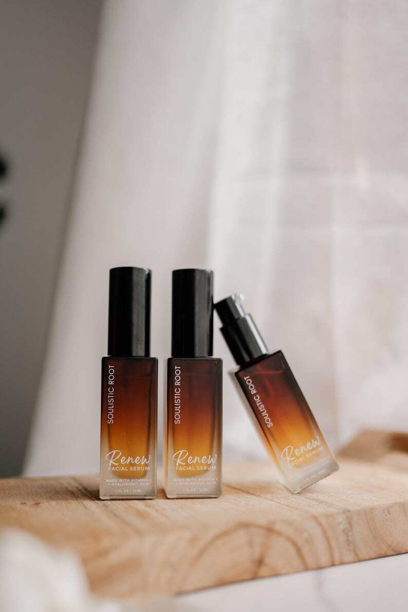 May include: Three bottles of Soulistic Root Renew Facial Serum on a wooden surface. The bottles have a gradient brown and clear design with black caps. The text "Renew Facial Serum" is visible on the bottles.
