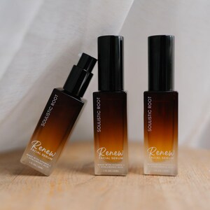 May include: Three brown and black facial serum bottles with the text "Soulistic Root" and "Renew Facial Serum". The bottles are arranged on a wooden surface with a white fabric backdrop.