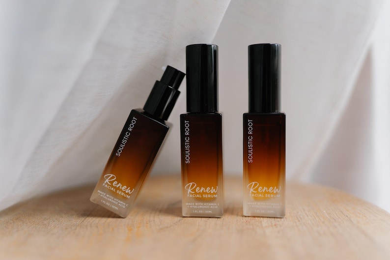 May include: Three brown and black facial serum bottles with the text "Soulistic Root" and "Renew Facial Serum". The bottles are arranged on a wooden surface with a white fabric backdrop.