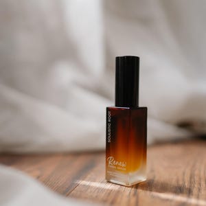 May include: A rectangular glass bottle with a gradient brown and yellow hue, topped with a black cap. The bottle has the text "SOULISTIC ROOT" and "Renew Facial Serum". The product is on a wooden surface.