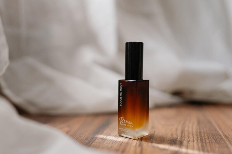 May include: A rectangular glass bottle with a gradient brown and yellow hue, topped with a black cap. The bottle has the text "SOULISTIC ROOT" and "Renew Facial Serum". The product is on a wooden surface.