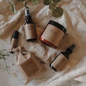 K&ouml;nnte beinhalten: Eine Sammlung von Bad- und K&ouml;rperprodukten, darunter ein Eukalyptus-Duschspray, ein entspannendes Kissenspray, ein Haferbad und ein belebendes Bad. Die Produkte werden auf einem hellbraunen Stoffhintergrund pr&auml;sentiert.