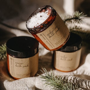 May include: Three amber glass jars with brown paper labels. The labels have white text that reads "revive bath soak", "seduction bath soak", and "cozy bath soak". The jars are sitting on a beige fabric surface with green pine needles.