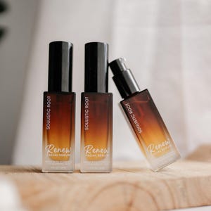 May include: Three bottles of facial serum on a wooden surface. The bottles are rectangular with a gradient color scheme of brown and orange. The labels read "Soulistic Root" and "Renew Facial Serum". One bottle is leaning.