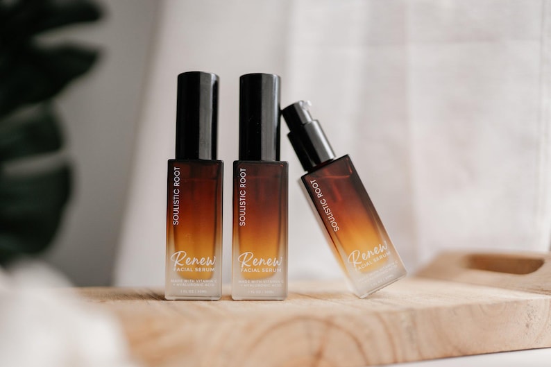 May include: Three bottles of facial serum on a wooden surface. The bottles are rectangular with a gradient color scheme of brown and orange. The labels read "Soulistic Root" and "Renew Facial Serum". One bottle is leaning.