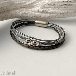 Puede incluir: Un brazalete de cuero gris con m&uacute;ltiples hebras y un dije de infinito plateado.