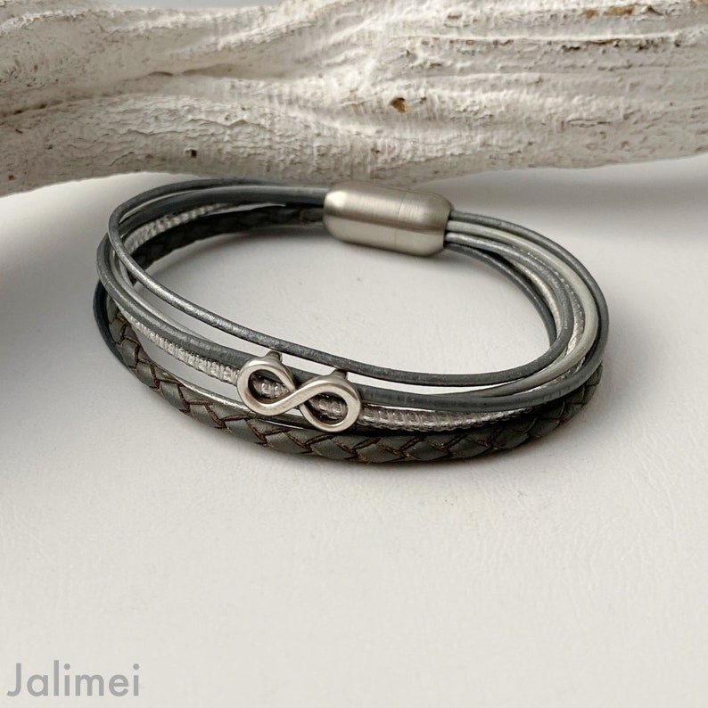 Puede incluir: Un brazalete de cuero gris con m&uacute;ltiples hebras y un dije de infinito plateado.
