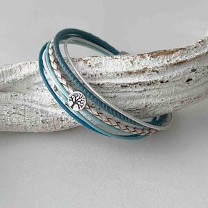 May include: A multi-strand bracelet with a silver charm featuring a tree of life design. The bracelet is made of braided leather and blue cord.