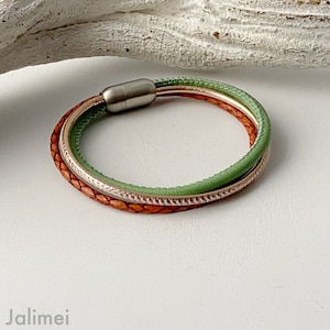 May include: A multi-strand bracelet featuring a magnetic clasp. The bracelet has three strands: a braided brown leather strand, a light green leather strand, and a thin, metallic gold-colored strand. The bracelet is designed to wrap around the wrist.