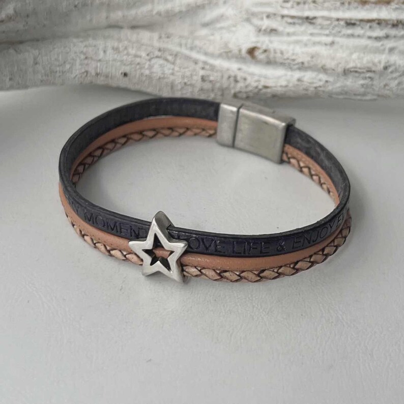 May include: A multi-layered leather bracelet with a silver star charm. The bracelet has a black leather strap with the words "Love Life & Enjoy Every Moment" embossed on it, and a tan leather strap with a braided design.