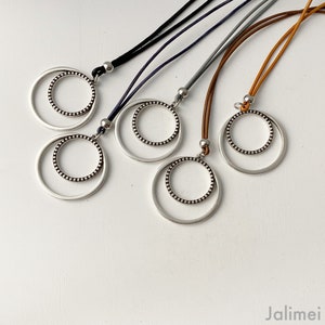 Può includere: Quattro collane con ciondoli a doppio cerchio in argento. Ogni ciondolo ha un cerchio più piccolo all'interno di un cerchio più grande. Le collane sono su cordini di diversi colori: nero, grigio, marrone e beige.