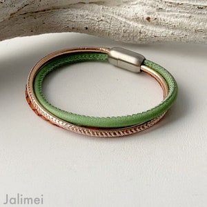 May include: A multi-strand bracelet featuring a light green leather cord, a copper-colored band with a textured design, and a rose gold band. The bracelet has a magnetic clasp. The jewelry is displayed on a white surface.