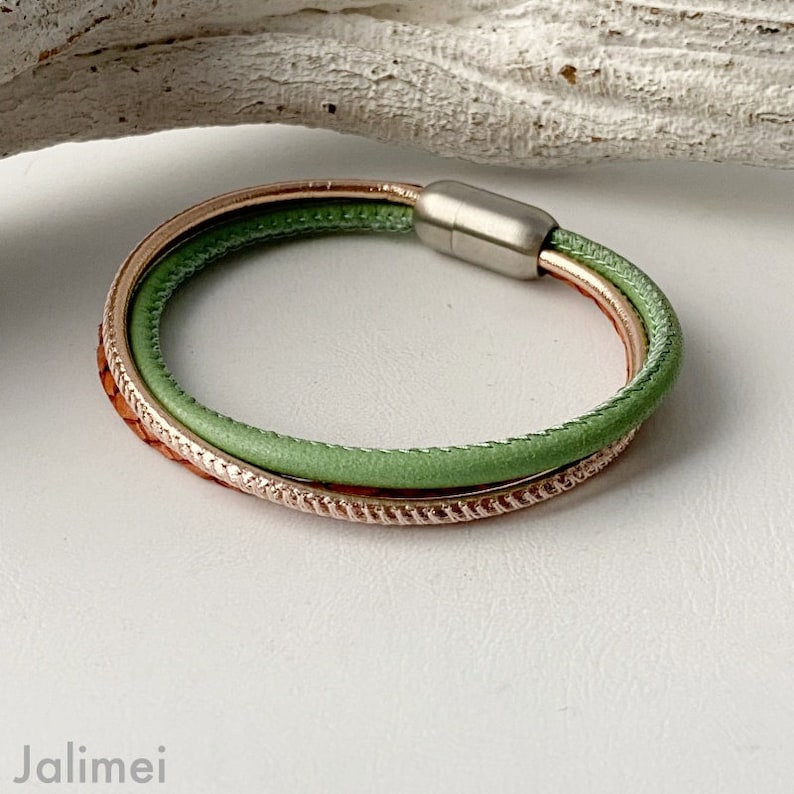 May include: A multi-strand bracelet featuring a light green leather cord, a copper-colored band with a textured design, and a rose gold band. The bracelet has a magnetic clasp. The jewelry is displayed on a white surface.