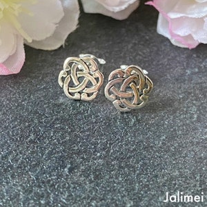 Könnte beinhalten: Ein Paar silberfarbene Ohrstecker. Jeder Ohrstecker hat ein keltisches Knotenmuster mit verschlungenen Linien, die eine kreisförmige Form bilden. Die Ohrringe werden auf einer dunkelgrauen Oberfläche mit weichen rosa und weißen Blumen im Hintergrund präsentiert.